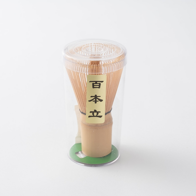 A 100-prong bamboo Chasen matcha whisk in a transparent cylindrical case with Japanese calligraphy on the label.