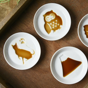 A collection of Kageasobi sauce plates filled with soy sauce to reveal the hidden designs of a cat, an owl, and Mount Fuji.

