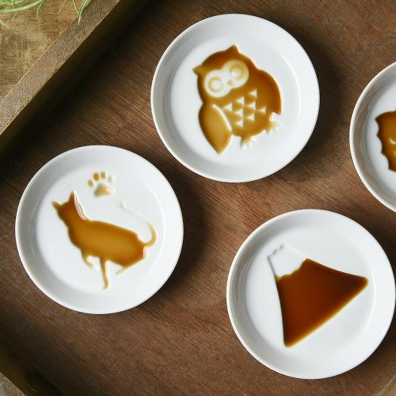 A collection of Kageasobi sauce plates filled with soy sauce to reveal the hidden designs of a cat, an owl, and Mount Fuji.

