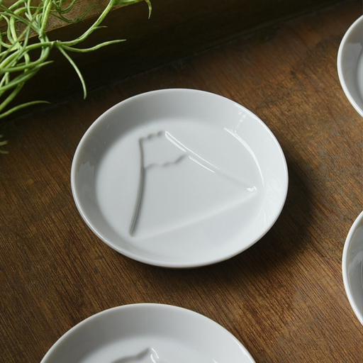 The Kageasobi Mount Fuji sauce plate, a small white porcelain dish with a subtle embossed design of the mountain, shown on a dark wood table.

