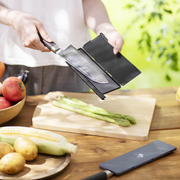 A person outdoors places a Damascus chef's knife into an open black Kai magnetic blade guard over a cutting board with fresh asparagus.