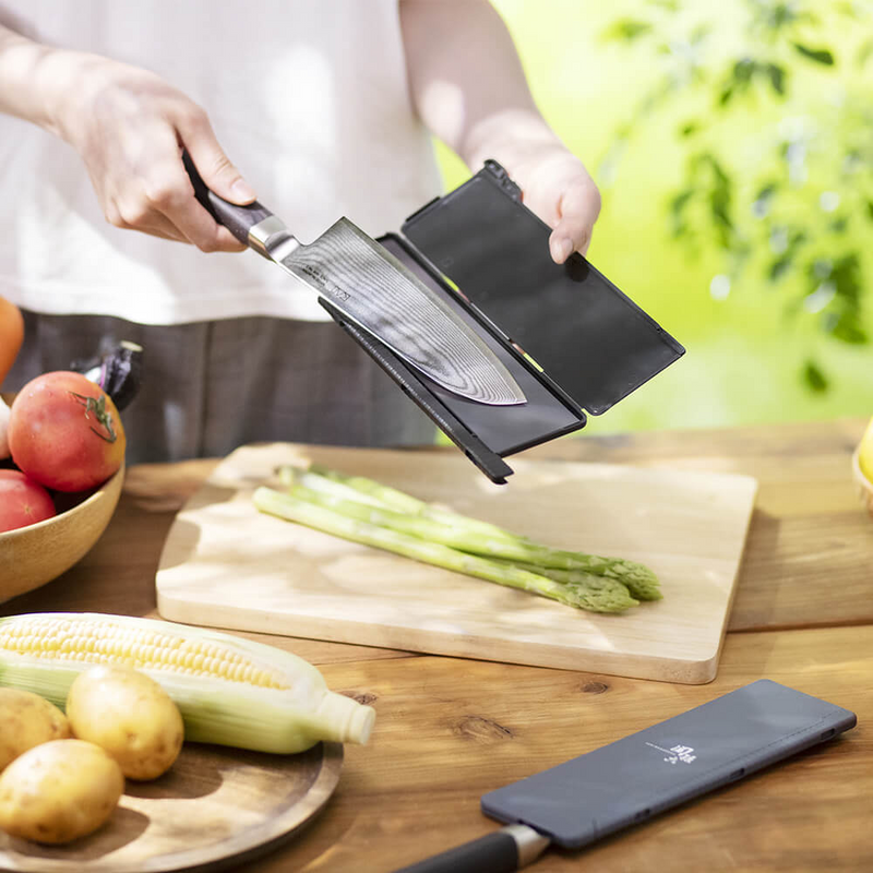 A person outdoors places a Damascus chef's knife into an open black Kai magnetic blade guard over a cutting board with fresh asparagus.