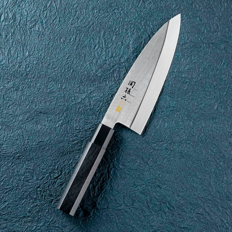 The Kai Seki Magoroku Premium Deba Knife (180mm) laid on a textured blue surface, showcasing its thick, single-bevel blade and traditional hexagonal composite wood handle.
