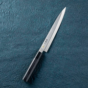 The Kai Seki Magoroku Sashimi Knife (210mm) laid on a textured blue surface, showcasing its long, slender, single-bevel blade designed for slicing sushi and sashimi.
