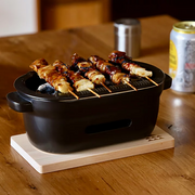 A black ceramic Kamoshika Douguten konro grill with yakitori skewers cooking over charcoal, placed on a wooden base on a dining table with a can of drink in the background.

