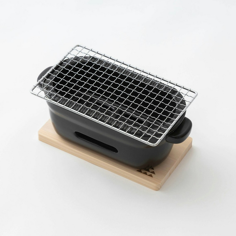 An angled top-down view of the black Kamoshika Douguten konro grill with its wire mesh on, placed on its wooden trivet against a white background.

