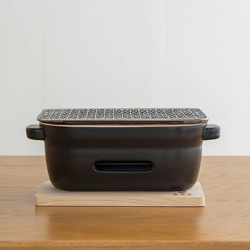 A straight side-on view of the Kamoshika Douguten konro grill, showing the ventilation slot and handles, sitting on its wooden base.