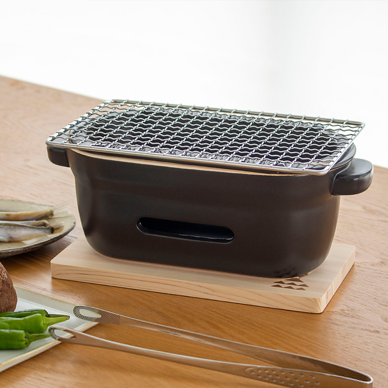 The Kamoshika Douguten konro grill on its wooden base, positioned on a light wood table next to tongs and a plate of food.

