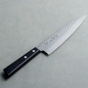 A Kaneyoshi Japanese chef knife with a hammered tsuchime blade finish and a black riveted handle, angled on a grey surface.