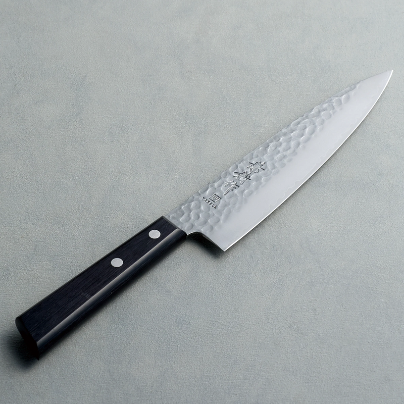 A Kaneyoshi Japanese chef knife with a hammered tsuchime blade finish and a black riveted handle, angled on a grey surface.