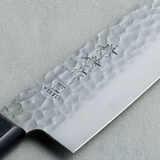 A close-up of the Kaneyoshi VG10 Santoku blade, detailing the hammered tsuchime finish, sharp edge, and Japanese kanji engraving.

