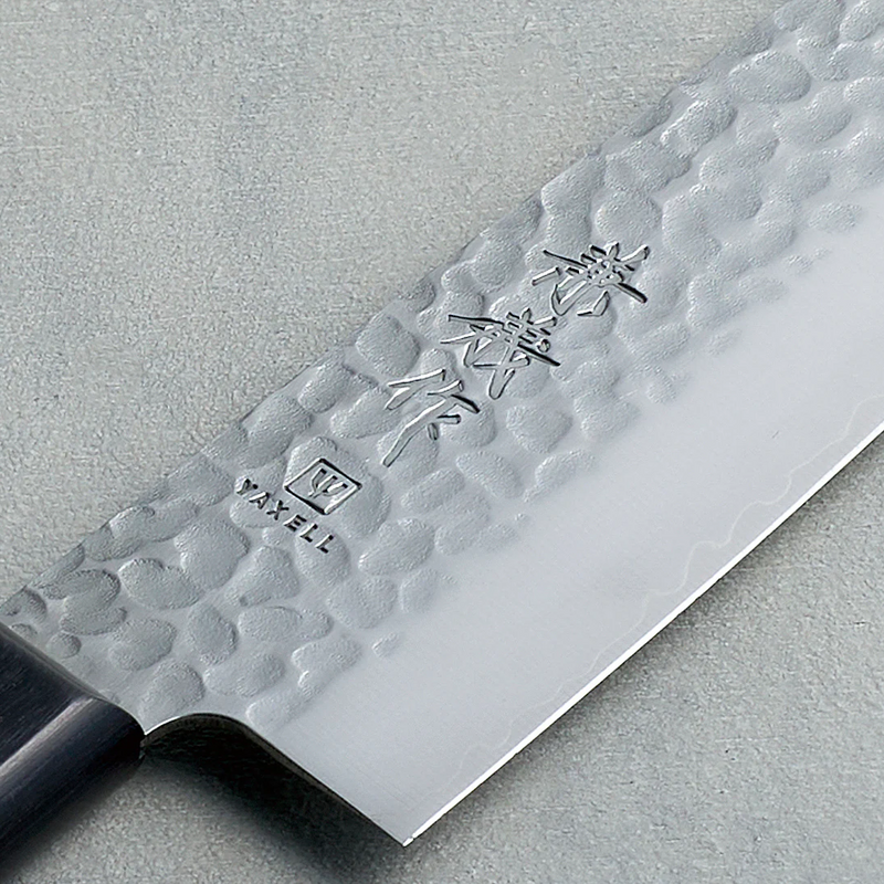 A close-up of the Kaneyoshi VG10 Santoku blade, detailing the hammered tsuchime finish, sharp edge, and Japanese kanji engraving.

