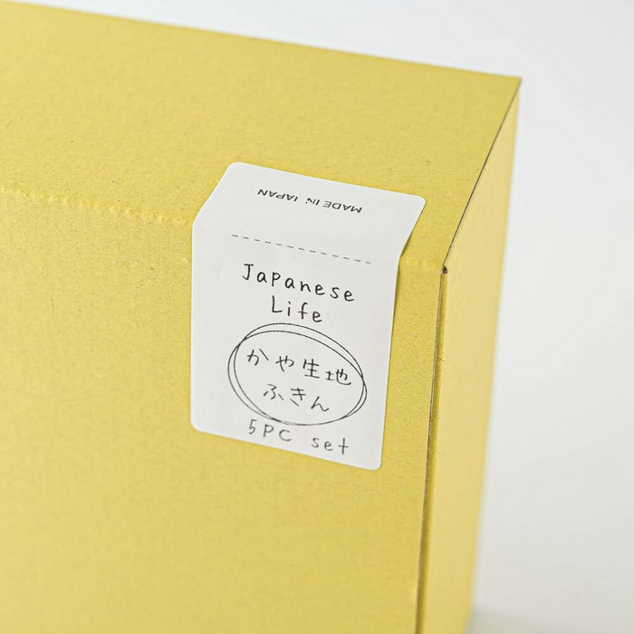 Close-up of a yellow gift box labeled 'Japanese Life' with a handwritten style sticker detailing 'かや生地ふきん, 5PC set' which translates to 'Kaya fabric cloths, 5 piece set.' The box texture is prominently displayed, emphasizing its simple, elegant packaging design.