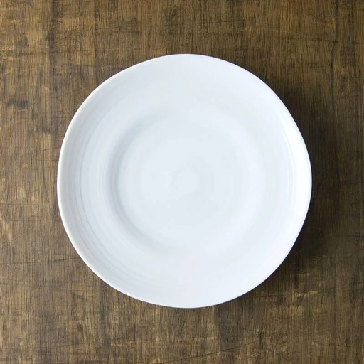 A top-down view of the white Kushime dinner plate, showing its subtle concentric lines, placed on a dark wood surface.

