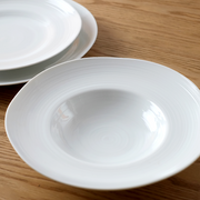 A top-down view of the white porcelain Kushime Dinner Hat Plate, showing its deep center and wide, textured rim, on a dark wood surface.