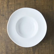 A top-down view of the white porcelain Kushime Dinner Hat Plate, showing its deep center and wide, textured rim, on a dark wood surface.
