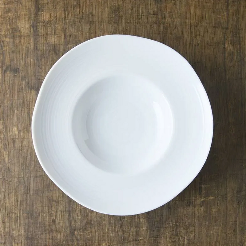 A top-down view of the white porcelain Kushime Dinner Hat Plate, showing its deep center and wide, textured rim, on a dark wood surface.

