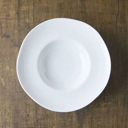 A top-down view of the white porcelain Kushime Dinner Hat Plate, showing its deep center and wide, textured rim, on a dark wood surface.
