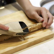 The Kyoto Katsugu forged peeler in use, smoothly peeling the skin from a long burdock root (gobo) on a wooden cutting board. 