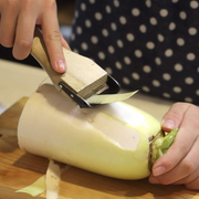 Close-up action shot of the sharp Kyoto Katsugu peeler effortlessly creating a long, thin peel from a large daikon radish. 
