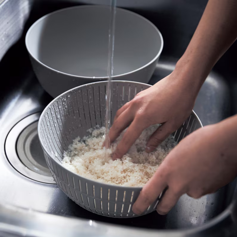 A person using their hands to wash rice in the grey Like-it colander under running water in a sink.