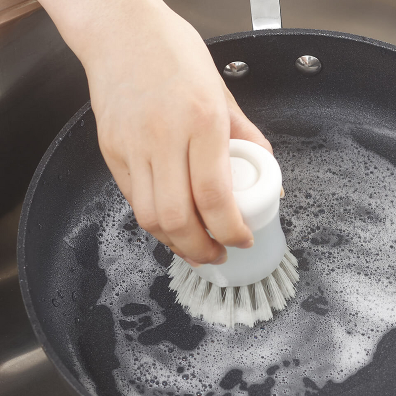 Scrubbing a pan with the Marna Antibacterial Detergent Dispenser Brush - A close-up of a hand using the Marna dispenser brush to scrub a pan filled with soapy water.
