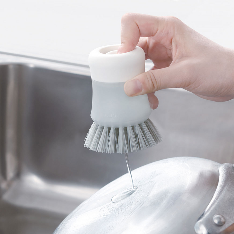 A hand using the Marna Antibacterial Detergent-Dispensing Brush, pressing the top button to release soap onto a pan.

