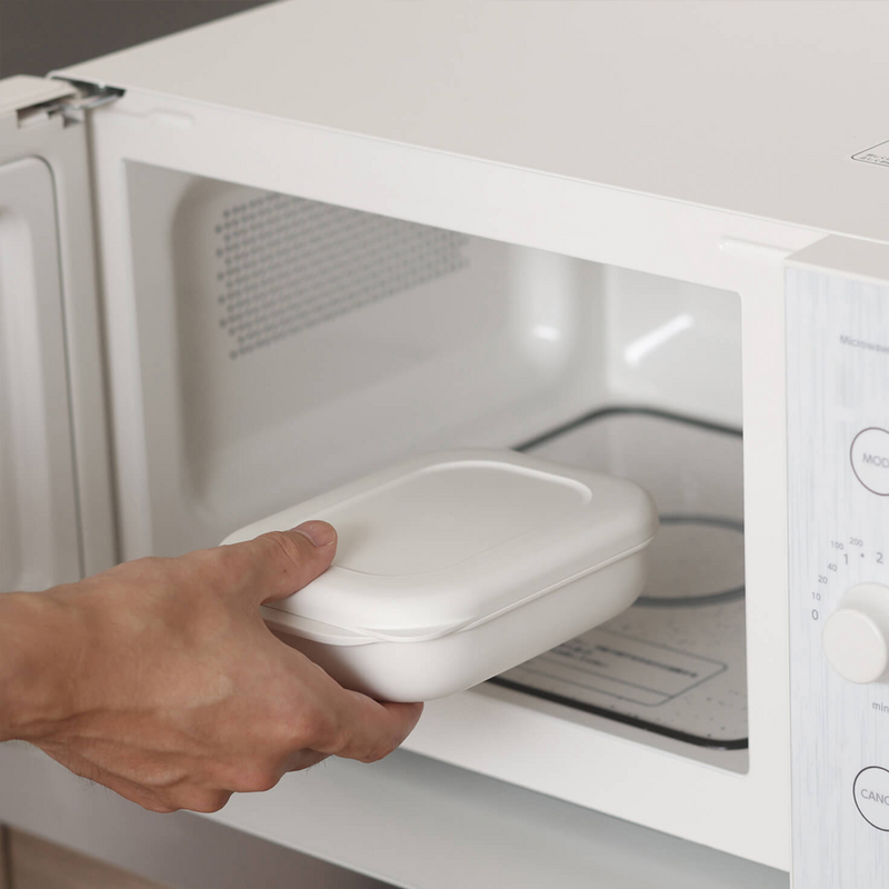 A hand placing the white Marna Kiwami Freeze & Microwave Rice Container into a microwave.