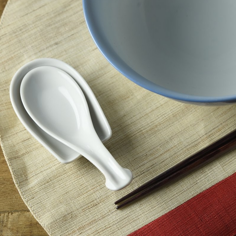 The Mino Yaki Classic Spoon and Spoon Rest Set in white, shown resting on a placemat next to a bowl.

