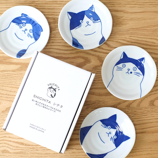 A set of five Shichita Mino Yaki cat side plates arranged on a wooden surface next to their white branded gift box.