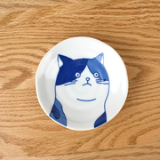 A top-down view of the Shichita Mino Yaki Cat Mike side plate, showing the cat's face with blue patches, resting on a wooden surface.
