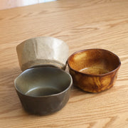A close-up of three unique bowls from the Mino Yaki Kuradashi gift set, showing their different rustic glazes and organic shapes on a wooden table.

