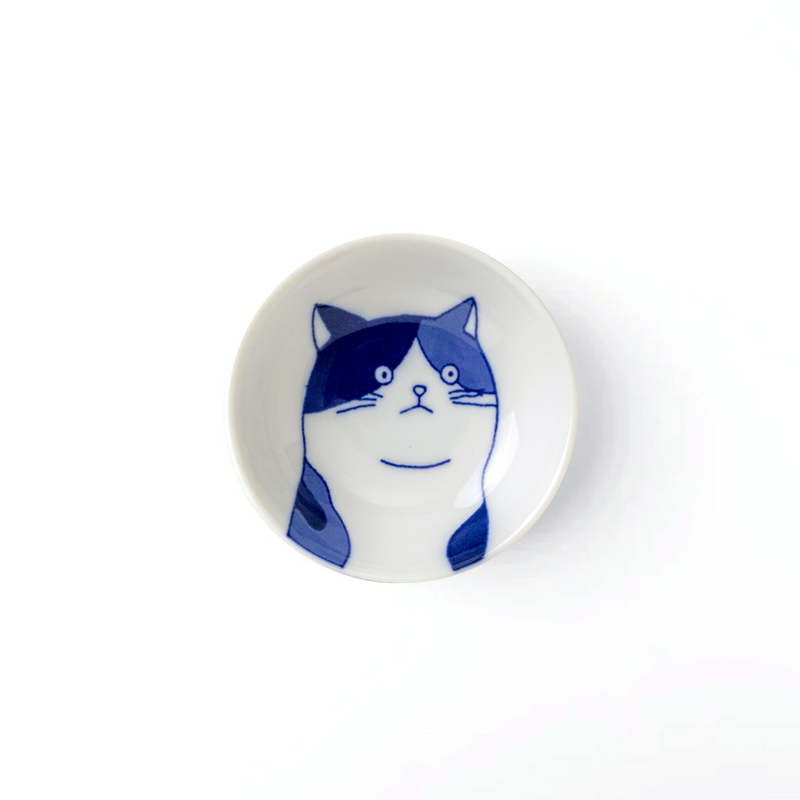 A ceramic side bowl with a depiction of a calm, contemplative cat's face on it, with wide, gentle eyes and a softly rounded shape, featuring a balanced blend of indigo markings on a crisp white background.