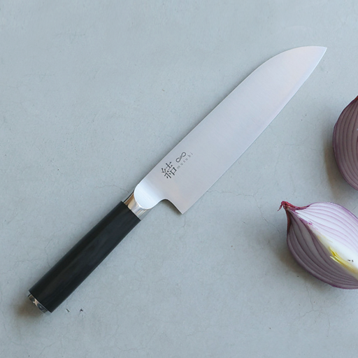A Mitsuboshi Musubi Santoku knife featuring a stainless steel blade with etched Japanese characters and a black pakkawood handle, displayed on a grey surface alongside sliced red onions.