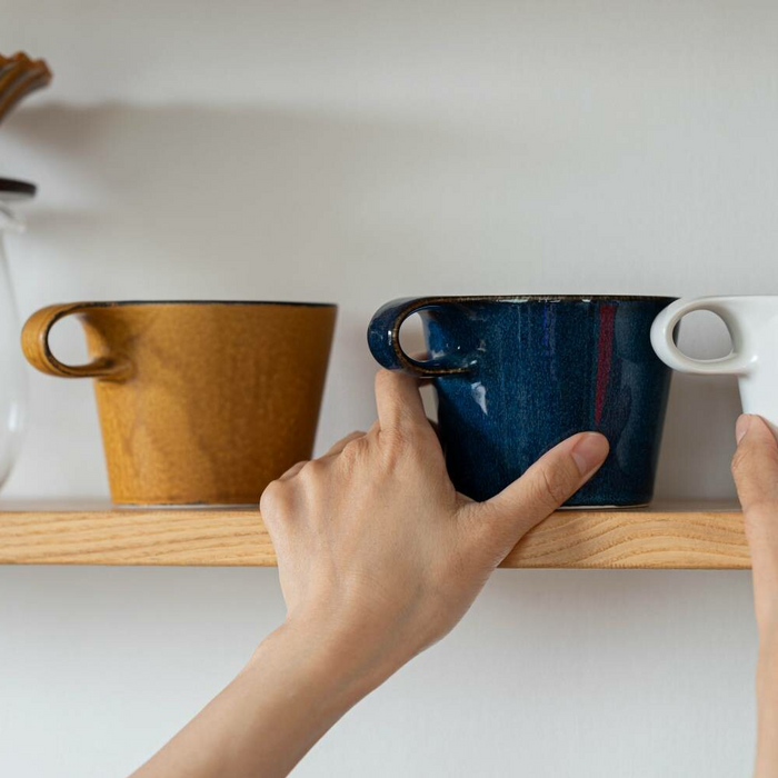 Person placing Miyama Metaphys Stamug in mustard colour on a shelf next to blue and white mugs.