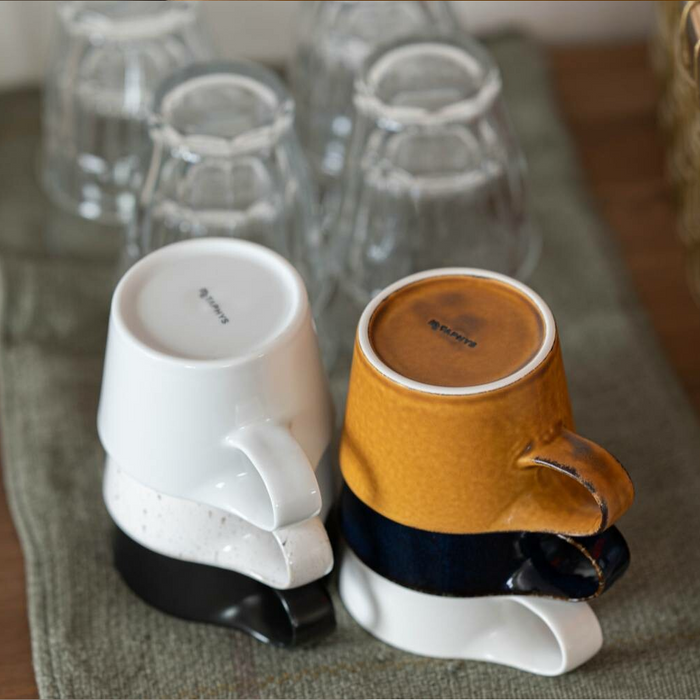 Several Miyama Bico Series stoneware mugs in different colors including white, mustard, and blue, stacked and displayed on a wooden shelf in a home setting.