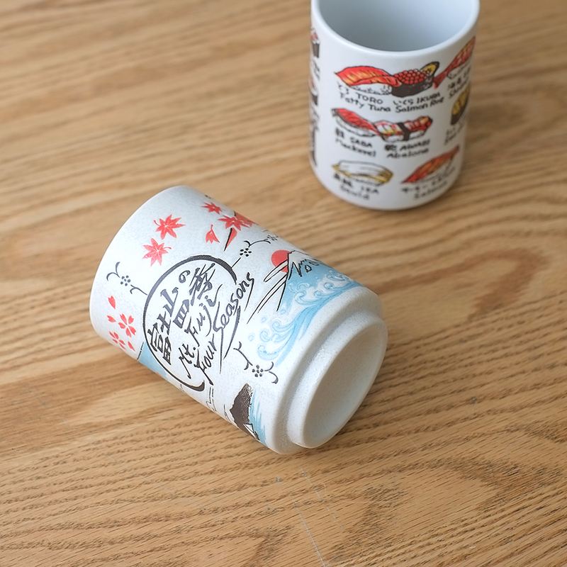 A cylindrical Japanese sushi teacup (yunomi) featuring illustrations of Mount Fuji across four seasons with cherry blossoms and Japanese calligraphy.