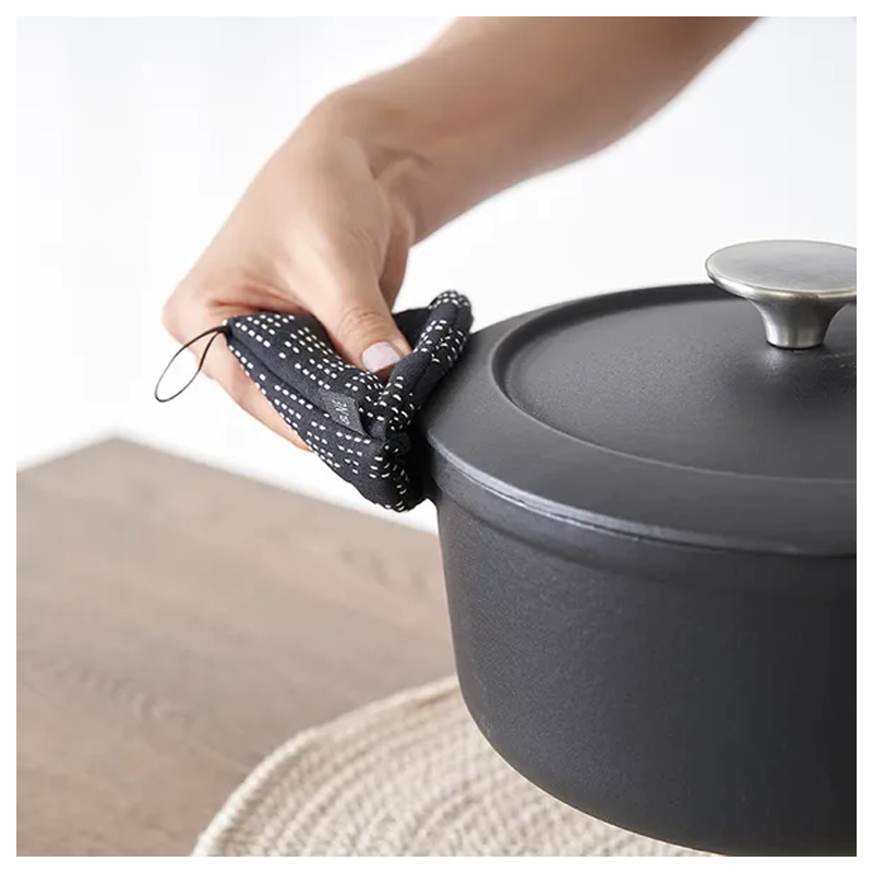 A black &NE pot holder with a dotted pattern being used to handle a dark gray pot on a neutral surface, with the &NE logo visible.