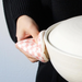 A person wearing a black sweater uses the pink checkered &NE pot holder to carry a large ceramic pot with a white lid, showcasing the pot holder's practical use in a kitchen setting.