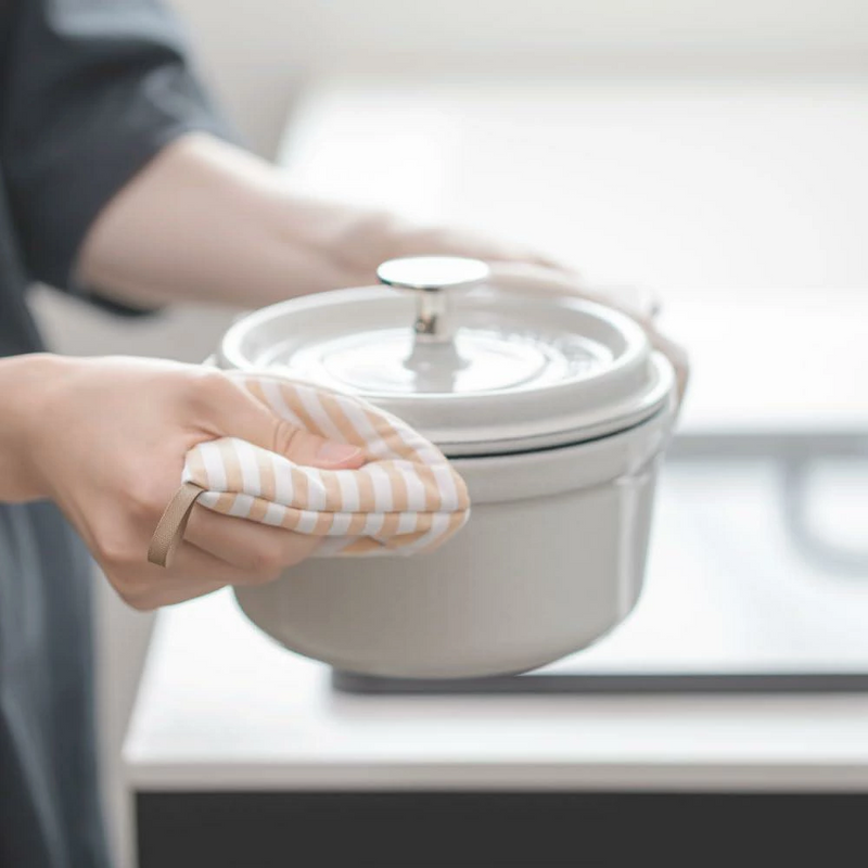 A person using the striped beige pot holder to grip the side handle of a hot enamel pot.