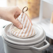 A hand using the &NE cone pot holder to safely lift the hot lid of a grey Staub cocotte by the knob.