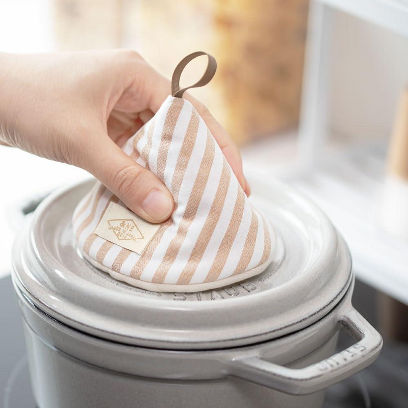 A hand using the &NE cone pot holder to safely lift the hot lid of a grey Staub cocotte by the knob.