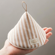 A close-up of a cone-shaped Japanese pot holder with beige and white stripes, held in a hand to show its size and leather hanging loop.