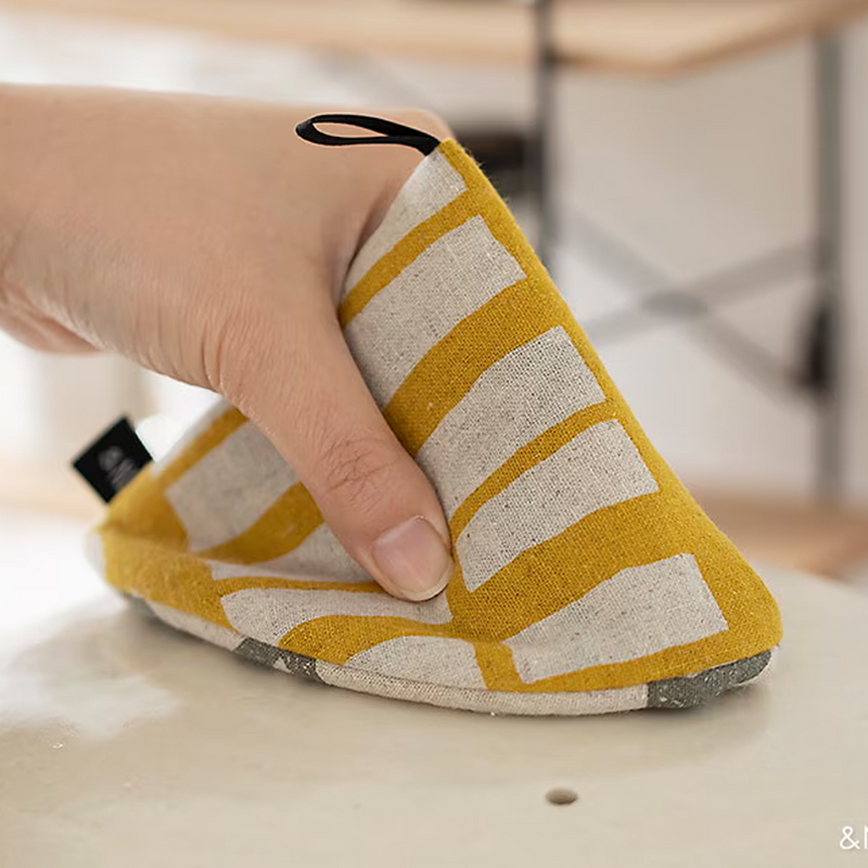 Hand gripping a NE Japanese yellow pot holder with white geometric pattern while holding a pot lid.

