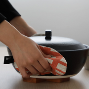 Hand holding a coral-patterned pot holder to support a donabe pot on a wooden table.

