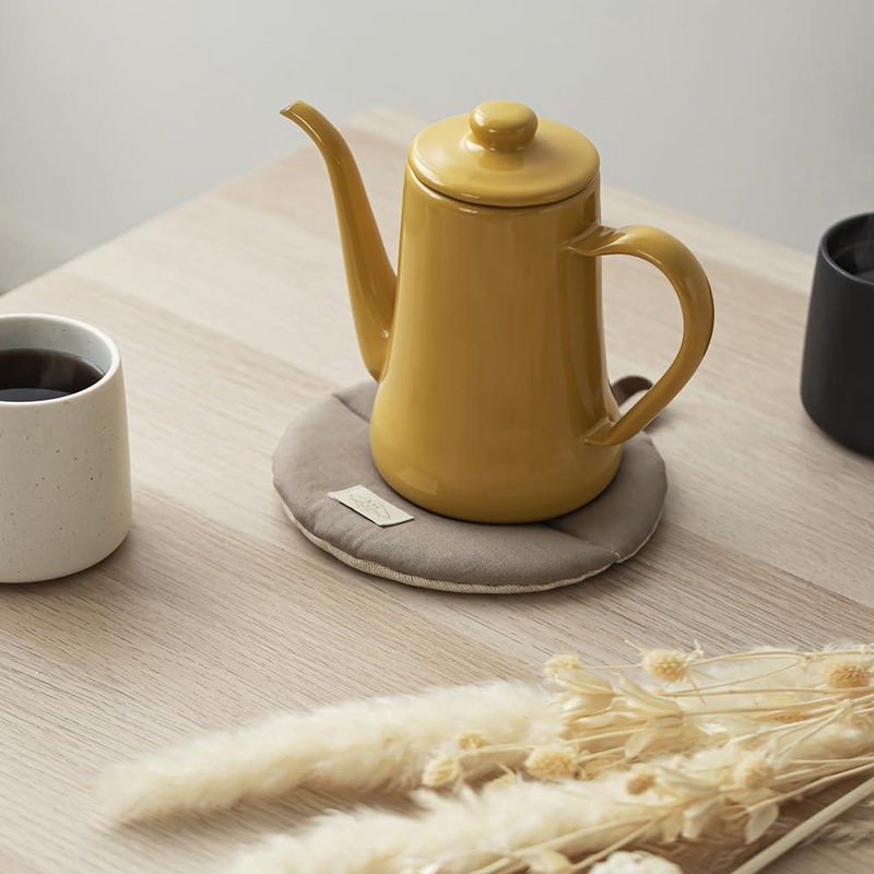 A yellow pour-over coffee kettle resting on the beige Manmaru table mat, used as a trivet to protect the wooden dining surface.