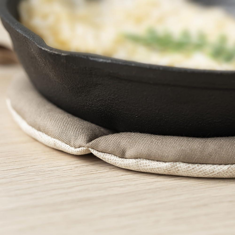 A close-up view of the edge of a round, natural beige fabric trivet supporting a black cast iron skillet on a wooden table.