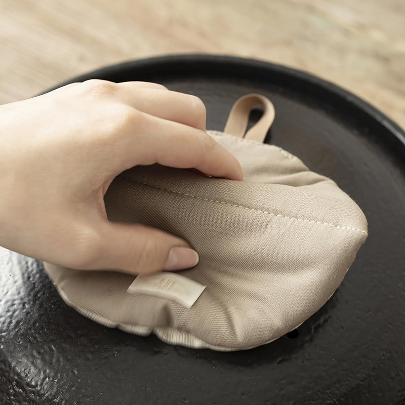 A hand using the folded beige Manmaru pot holder to safely grip the hot handle of a black cast iron pot lid.