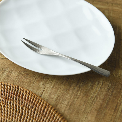 Nagomi Hammered Style two-pronged stainless steel fruit fork with tsuchime texture, resting on a white dimpled plate on a wooden table with a rattan placemat
