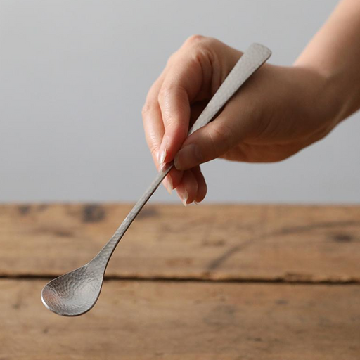 Nagomi Hammered Style stainless steel soda spoon with long slender handle and tsuchime texture, held in a hand above a rustic wooden table
